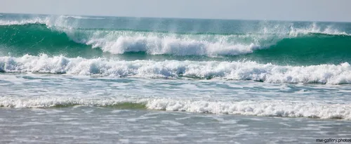 This landscape photograph captures waves breaking along the coast at Sennen Cove in Cornwall, United Kingdom. Taken in the afternoon during the spring season, the image shows the natural movement of water rolling onto the beach, with sea spray visible above the cresting waves. The setting highlights the beauty of nature at the Cornish coast, showcasing the dynamic ocean environment typical of this region. The foreground includes the shallow water and foaming surf characteristic of beaches in Cornwall.
